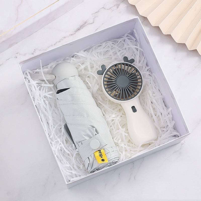 A gift box on a marble surface containing a folded grey umbrella and a handheld white fan with bear ears, nestled in white paper shreds.