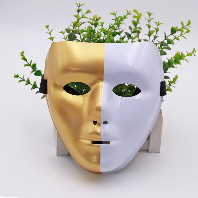 A half-gold, half-white mask with eye and mouth openings, set against a background of green plants emerging from behind, creating a striking visual contrast.