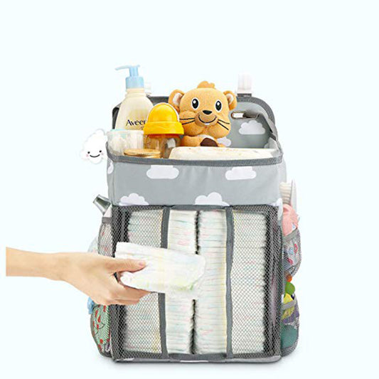 Gray diaper caddy with cloud pattern, filled with baby essentials including diapers, wipes, lotion, a toy lion, and more. A hand reaches for a diaper from the front pocket.