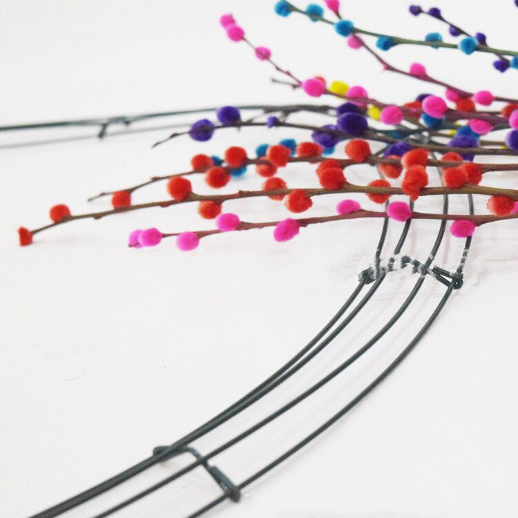 Colorful pom-pom branches arranged on a wire wreath form against a plain white background, featuring red, pink, purple, blue, and yellow fluffy balls.