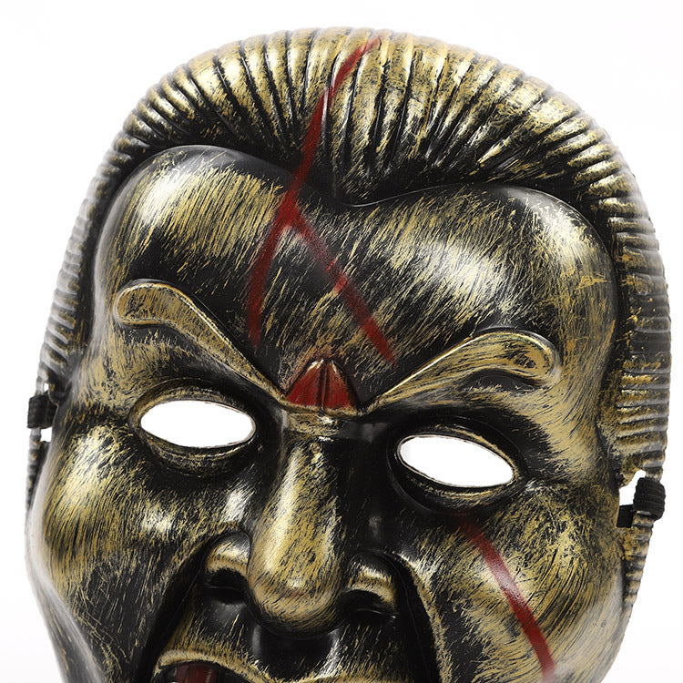 A black and gold metallic mask with exaggerated facial features and red slashes. It has prominent brows, hollow eyes, and detailed texture resembling aged metal.