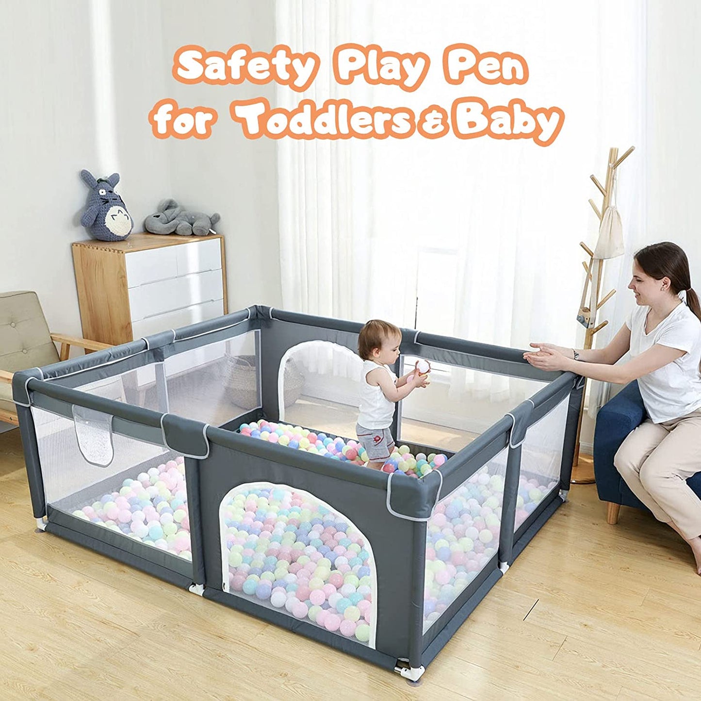 A toddler plays in a gray safety playpen filled with colorful balls. An adult sits nearby in a living room setting with a plush toy on a cabinet and a coat rack in the corner.