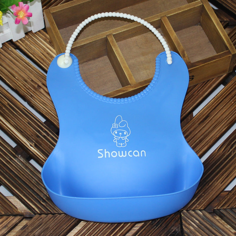 Blue silicone bib with a scoop pocket and a cute character design, labeled "Showcan." Features a beaded adjustable strap, placed on a wooden surface with flowers nearby.