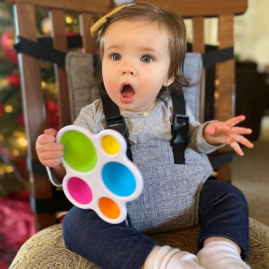 A wide-eyed baby with a surprised expression sits strapped to a high chair, holding a colorful toy with multicolored bubbles. A Christmas tree is visible in the background.