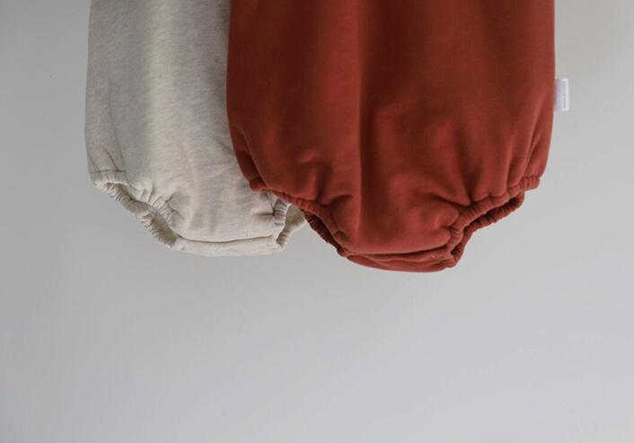 Two baby rompers hanging side by side against a neutral background. One is light beige, and the other is a warm rust color, both with elastic leg openings.