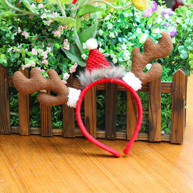 A reindeer antler headband with a red Santa hat, featuring white trim and faux fur, is placed on a wooden surface in front of a small wooden fence with green and pink flowers.