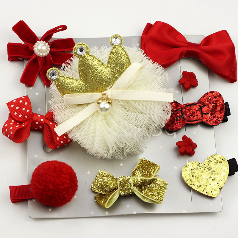 A collection of hair accessories includes red bows, a gold glitter heart, a gold bow, a red pom-pom, a glittery crown on tulle, and flower-shaped clips on a white board.
