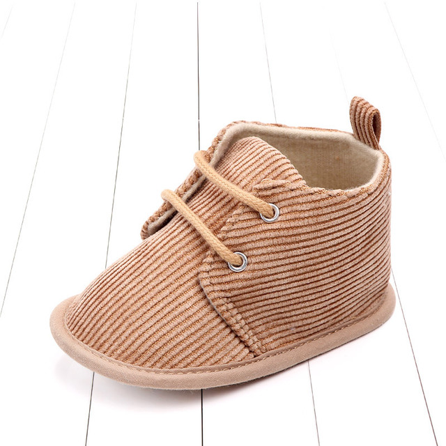 Tan corduroy baby shoe with lace-up design on a white wooden floor. The shoe features a soft interior and a loop at the back, suitable for infants.