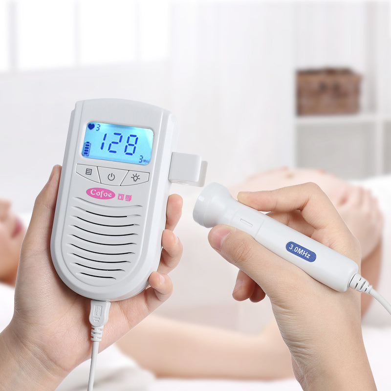A person holds a handheld fetal doppler device displaying "128" on the screen, with a probe in the other hand, used for monitoring fetal heartbeat in a clinical setting.
