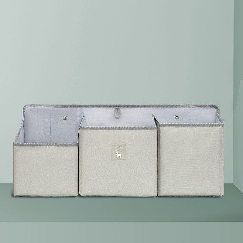 Three light gray fabric storage bins with a smooth finish are arranged side by side against a matching background. The middle bin features a small decorative label.