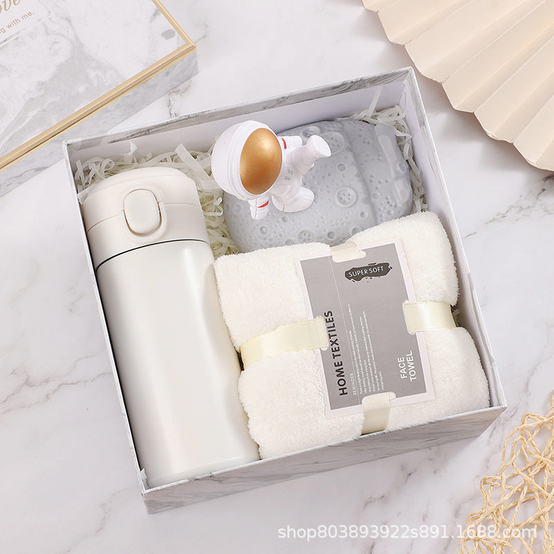 Gift box with a white insulated tumbler, a rolled white towel labeled "SUPER SOFT HOME TEXTILES," and a small astronaut figurine on a gray moon-shaped item.