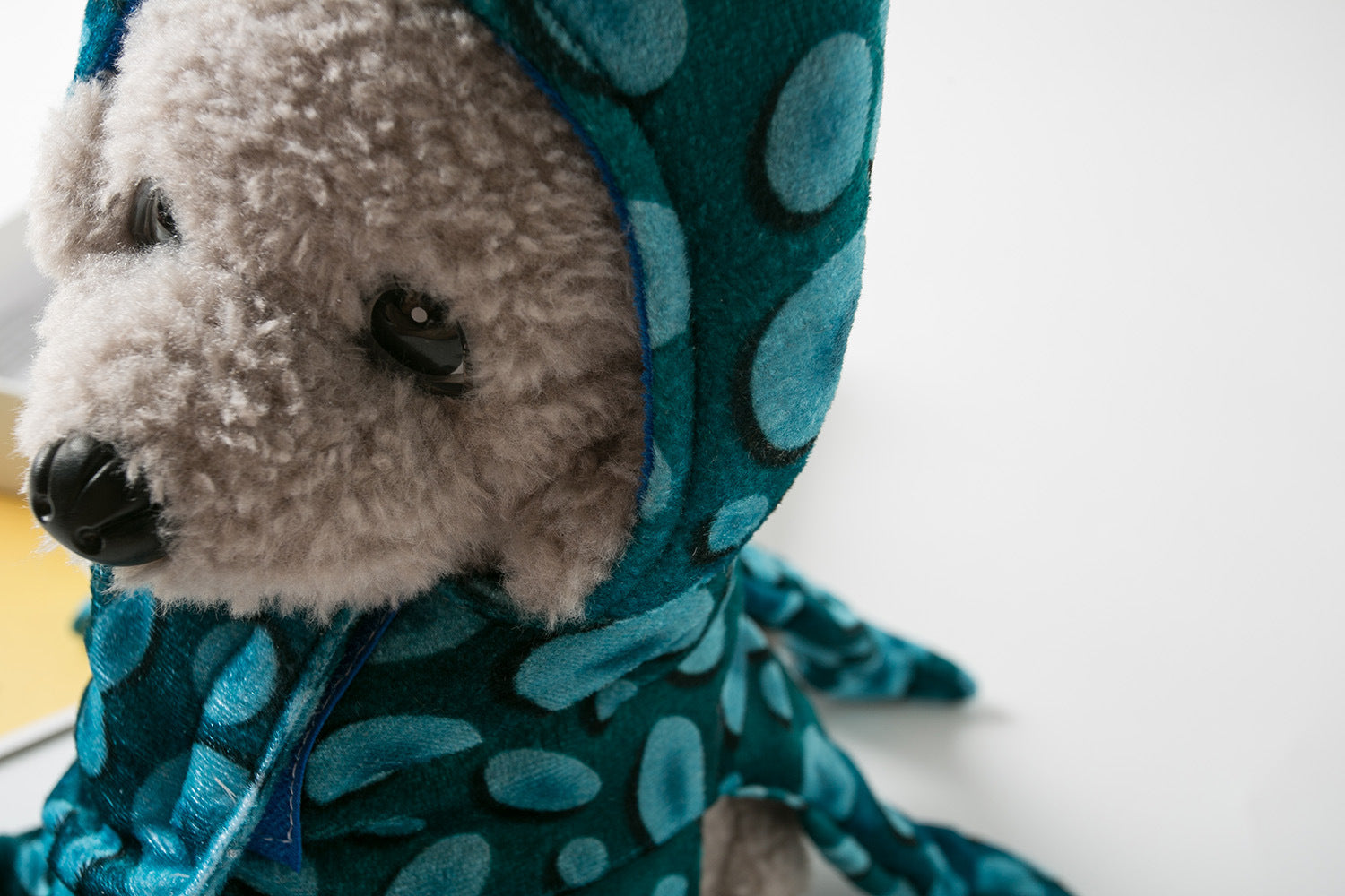 Close-up of a plush toy dog wearing a blue polka-dot hooded outfit. The fur is curly and beige. The background is softly blurred and white.
