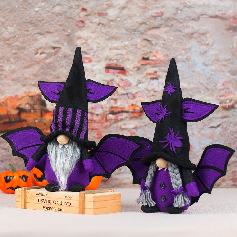 Two plush gnome figurines in purple witch costumes with bat wings stand on a wooden box. The left gnome has a striped hat and long beard, while the right one has a spider-adorned hat and braided hair. Small orange pumpkins are in the background against a textured brick wall.