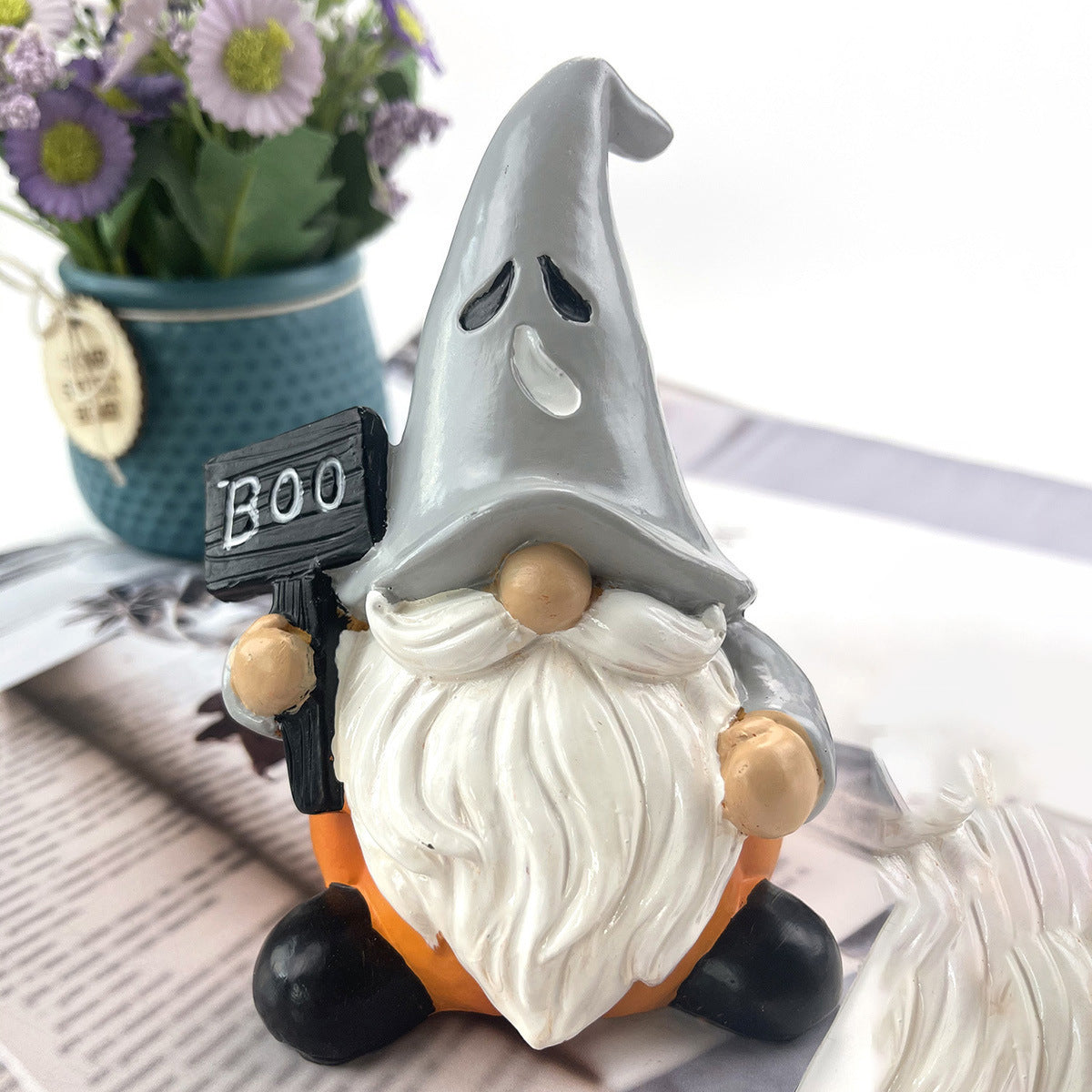 A gnome with a gray hat and white beard holds a "Boo" sign. It sits on a table with an orange pumpkin base. A pot of flowers is in the blurred background.