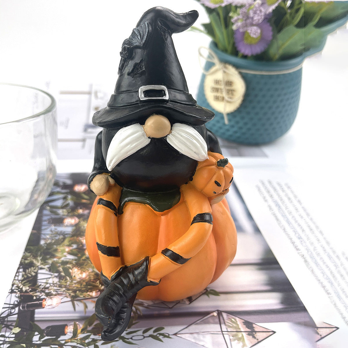 A whimsical gnome in a black witch hat and white mustache sits on an orange pumpkin, holding a tiny pumpkin. A blue pot with flowers is blurred in the background.