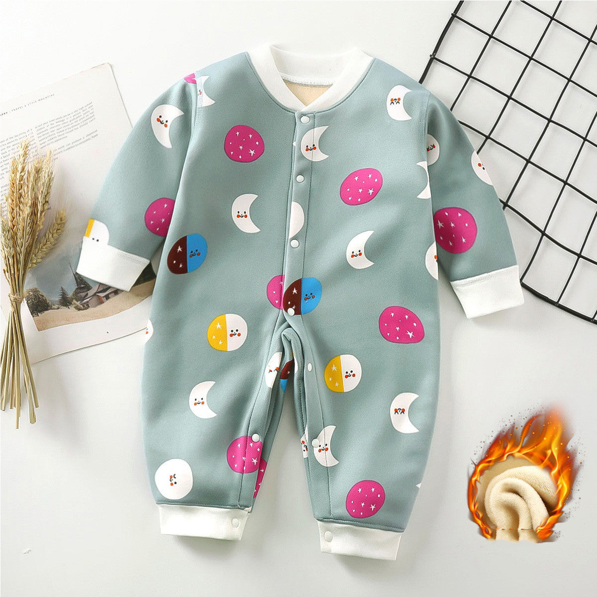 Baby romper with colorful moon and dot patterns on gray fabric, featuring white cuffs and snap buttons. Nearby, a small bunch of wheat and a grid pattern are visible.