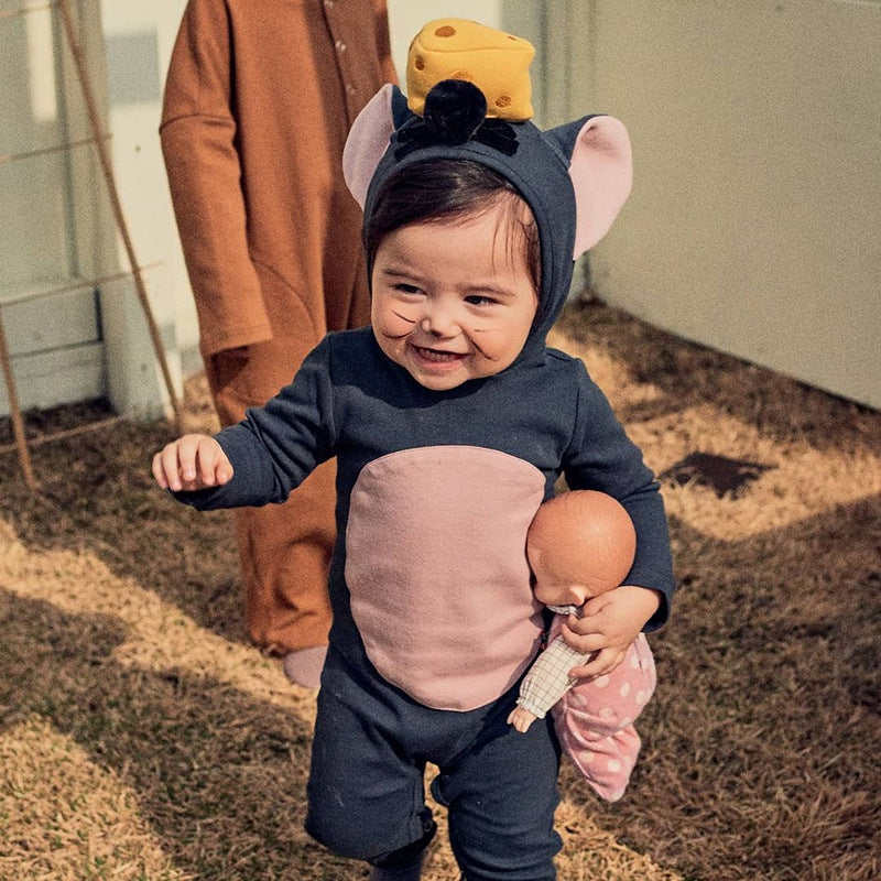 A smiling toddler in a mouse costume with pink ears and a cheese hat holds a doll while walking on grass. Another child in a brown outfit stands nearby.