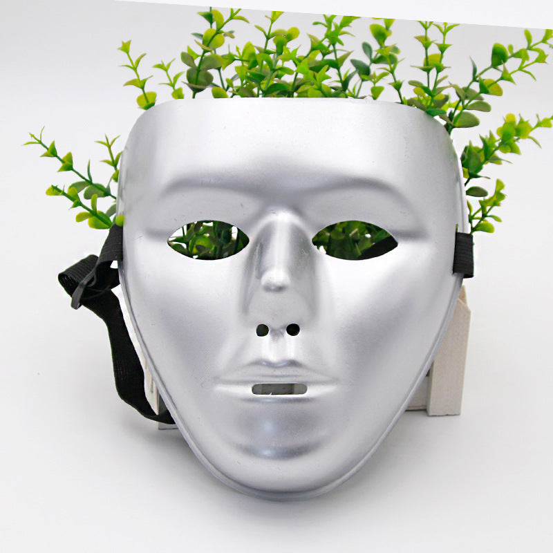 A silver metallic mask with black straps is positioned upright in front of a backdrop of green leafy plants, creating an intriguing visual contrast.