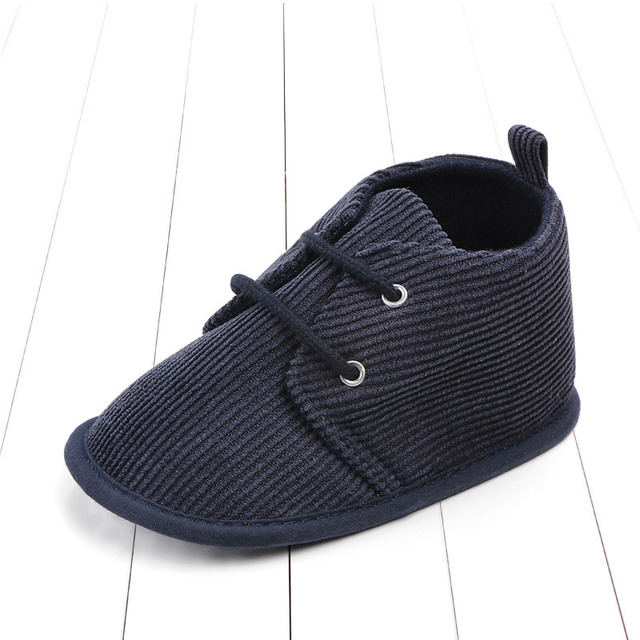 A navy blue ribbed fabric baby bootie with a soft sole and two eyelets for laces, displayed on a white wooden floor.