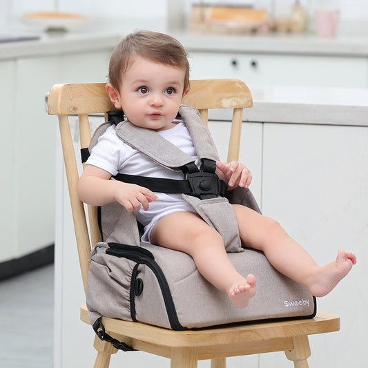 Baby seated in a light beige portable high chair with safety straps, on a wooden chair in a bright kitchen setting, wearing a white onesie and gazing to the left.