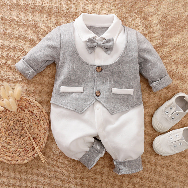 A baby outfit with a gray bow tie and vest over a white onesie, paired with matching gray cuffs. Nearby are white baby shoes and a woven mat with decorative grasses.