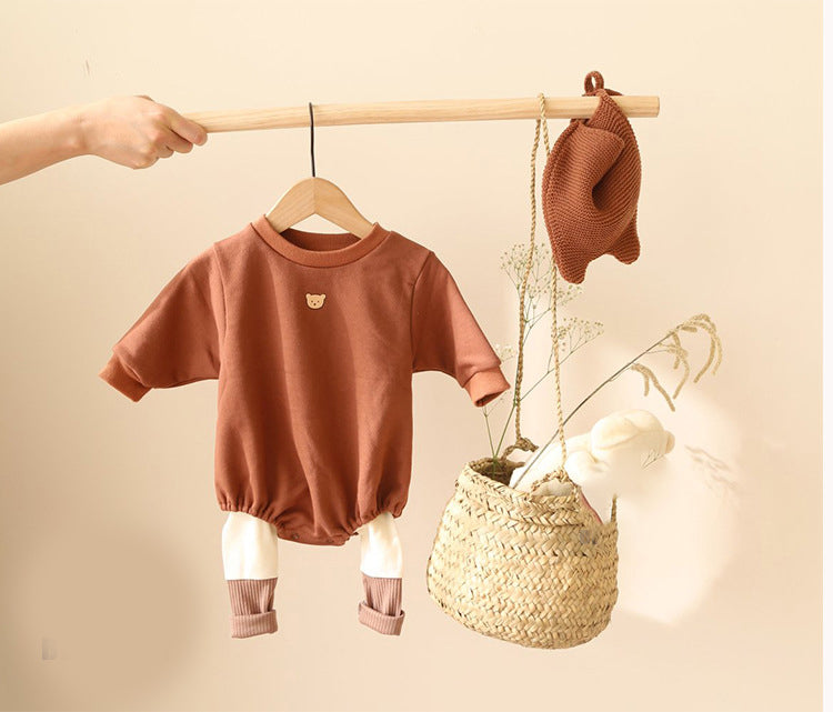 A hand holds a wooden rod displaying a brown baby romper with white leg cuffs and bear emblem. Nearby, a straw basket contains a bunny toy and beige knit hat.