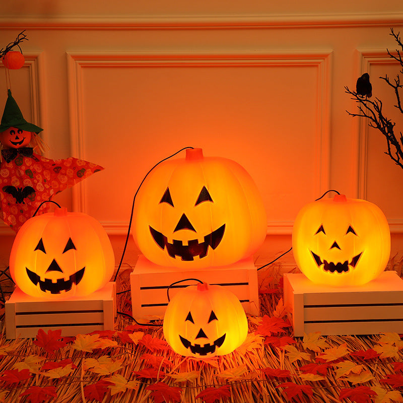 Four glowing jack-o'-lanterns with carved faces sit on white crates surrounded by autumn leaves. A small scarecrow and a bare branch add festive Halloween ambience.