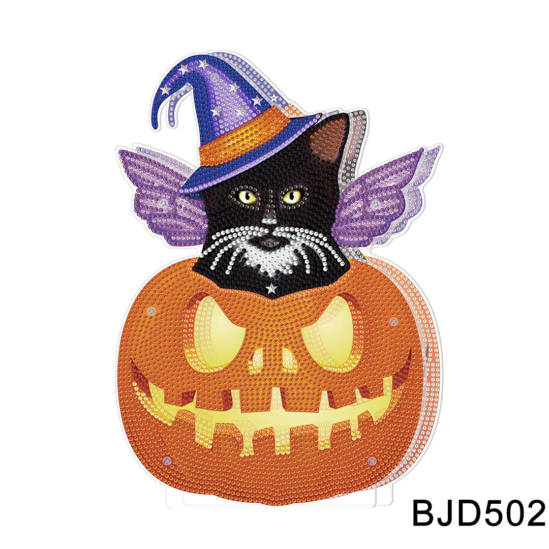 A decorative art piece featuring a black cat with a purple witch hat and wings, sitting inside a glowing jack-o'-lantern. The design is embellished with colorful gems.