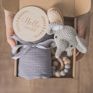 Close-up of a gift box containing a crocheted gray elephant toy, a soft gray fabric tied with a ribbon, a wooden teether, and a round plaque with "Hello world" engraved.