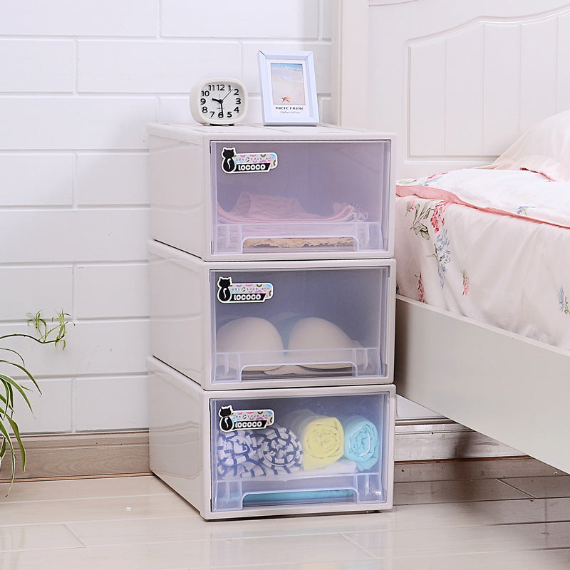 Three white storage drawers by a bed, holding neatly folded clothes and towels. A clock and photo frame sit on top. The wall and floor are white with light wood accents.