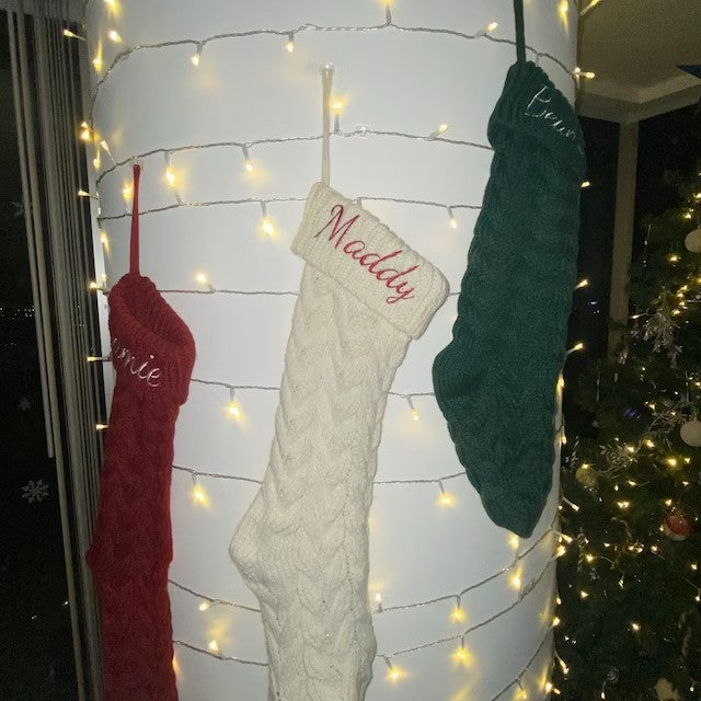 Three knit Christmas stockings in red, white, and green hang on a lit white post, embroidered with names. A decorated Christmas tree is visible in the background.