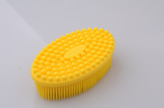 A yellow rubber scrub brush with an oval shape, covered in rounded bumps on top and bristles on the bottom, lying on a white surface.