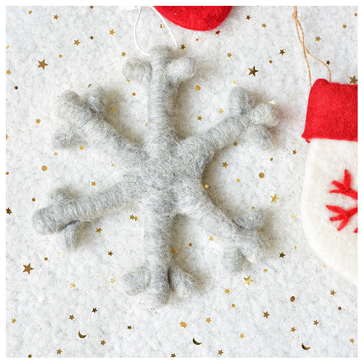 A felt grey snowflake ornament resting on a white, star-patterned background with a partial view of a red and white stocking ornament.