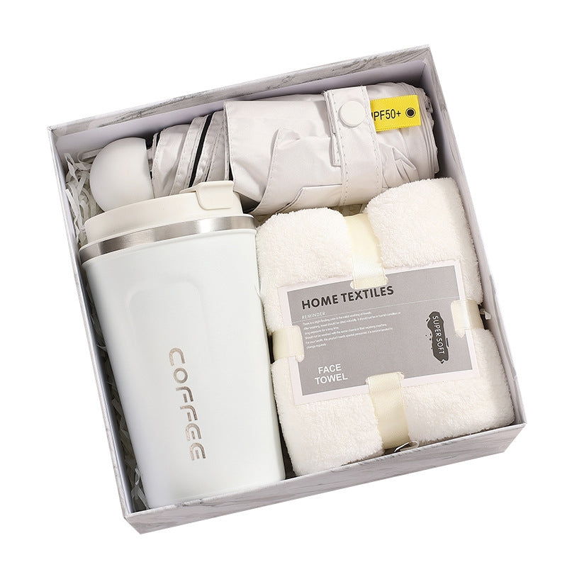 Gift box containing a white insulated mug labeled "COFFEE," a rolled gray umbrella with a "50+" tag, and a soft white face towel labeled "HOME TEXTILES."