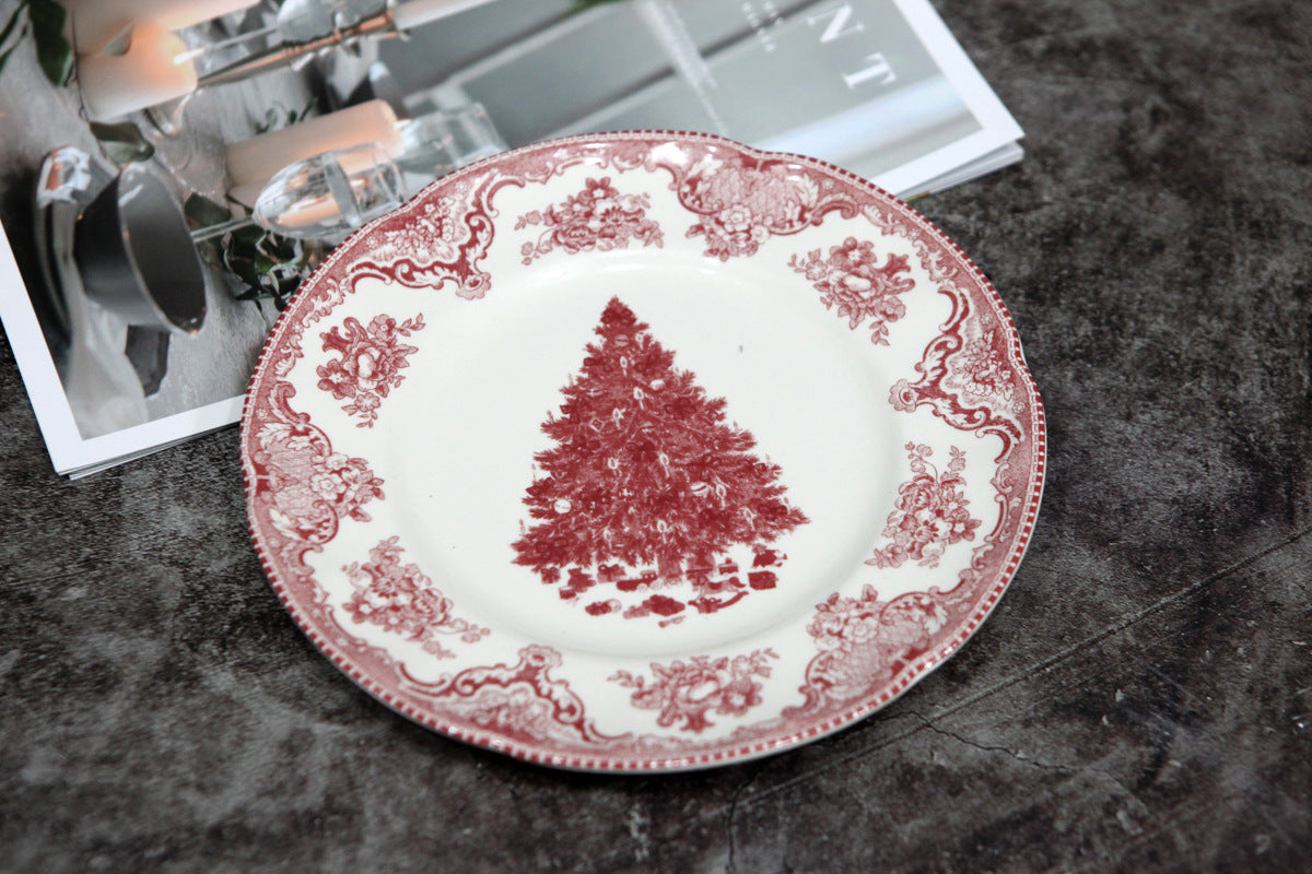 A decorative plate with a red Christmas tree in the center, surrounded by intricate floral patterns on the rim, rests on a textured surface beside an open magazine with candles.