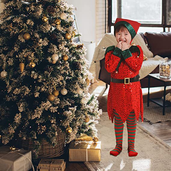 A child dressed as a festive elf stands excitedly beside a decorated Christmas tree with gold and white ornaments, in a cozy living room with gifts underneath the tree.