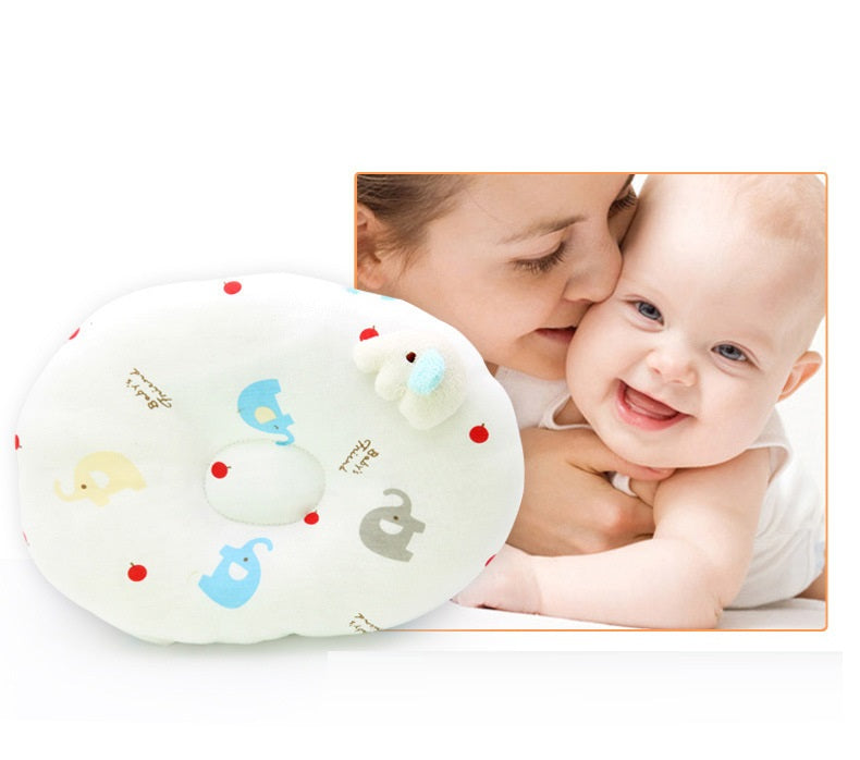 A smiling baby held by a woman, next to a colorful baby pillow with elephant and apple designs, featuring a small plush toy on top.