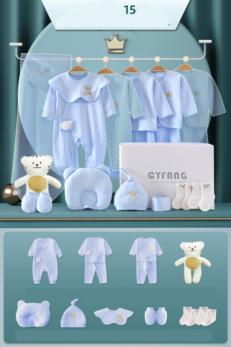 Blue baby clothing set with onesies, jackets, pants, hat, socks, bib, pillow, and teddy bear toy. Items are hung or placed on a display with a white gift box.