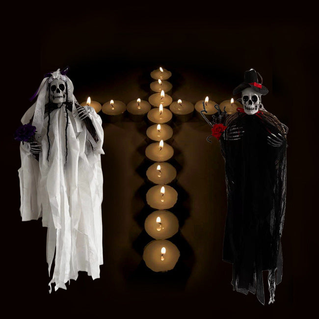 Two skeleton figures dressed as a bride and groom, each holding flowers, stand on either side of a cross made from lit candles against a dark background. The bride wears a white dress and veil, while the groom is in black with a hat and red rose.