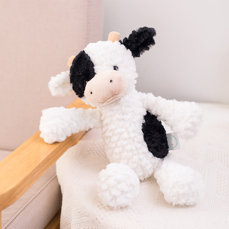A fluffy black and white cow plush toy with a soft pink snout sits on a cozy chair with a textured blanket draped over it.