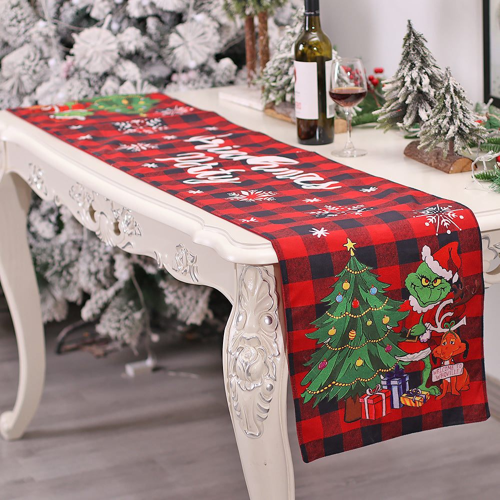 Festive table with a red plaid runner featuring a green character, Christmas tree, and presents. Snowflake accents, wine bottle, and frosted mini trees complete the holiday decor.