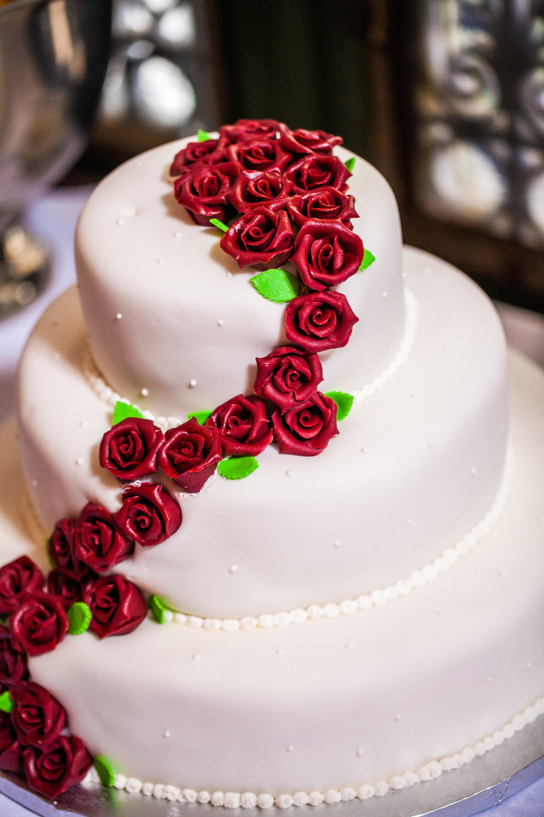 Three-tiered white cake adorned with cascading red sugar roses and green leaves, featuring small pearl-like accents along each layer's edge.