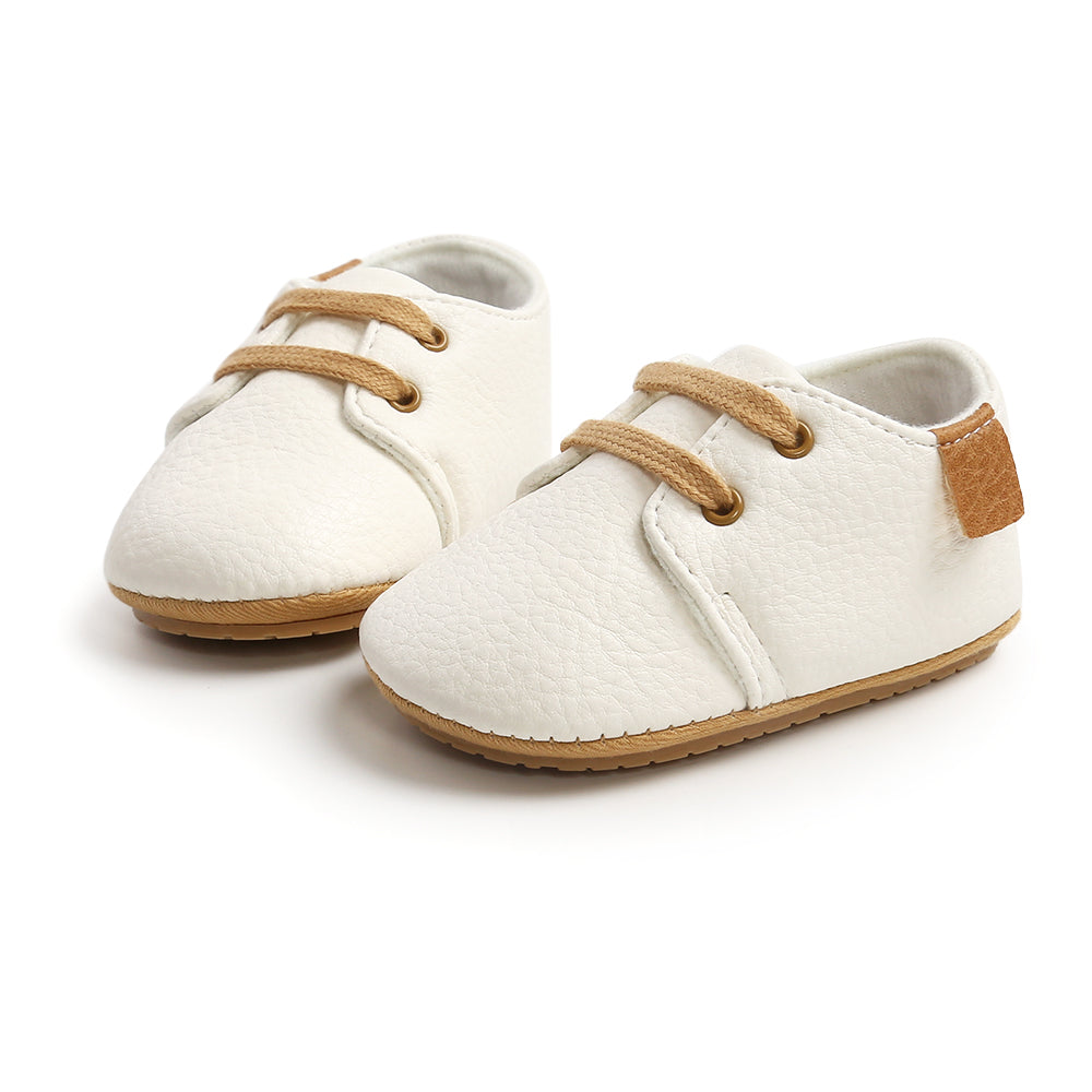 White baby shoes with tan laces and brown soles, featuring a faux leather texture and a small brown patch on the heel. The shoes are arranged side by side on a white background.