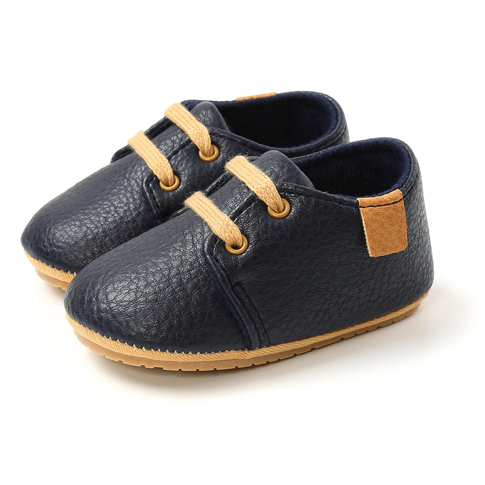 Navy blue baby shoes with tan laces and soles, featuring a textured surface and a small tan patch on the heel, displayed against a white background.