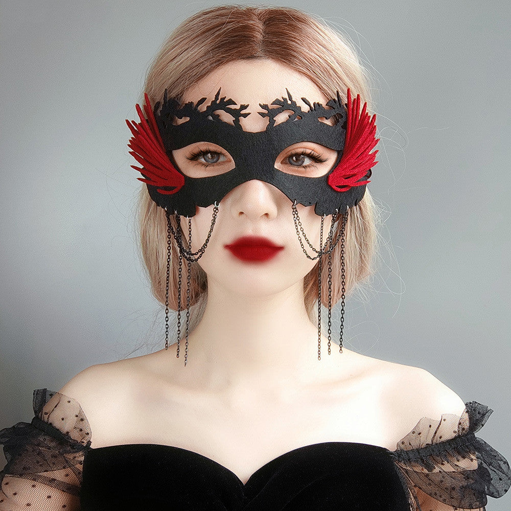 Woman in a black lace mask with red accents and chain details, wearing a sheer black dress with puffed sleeves. Her lips are painted dark red, and her hair is light blonde.