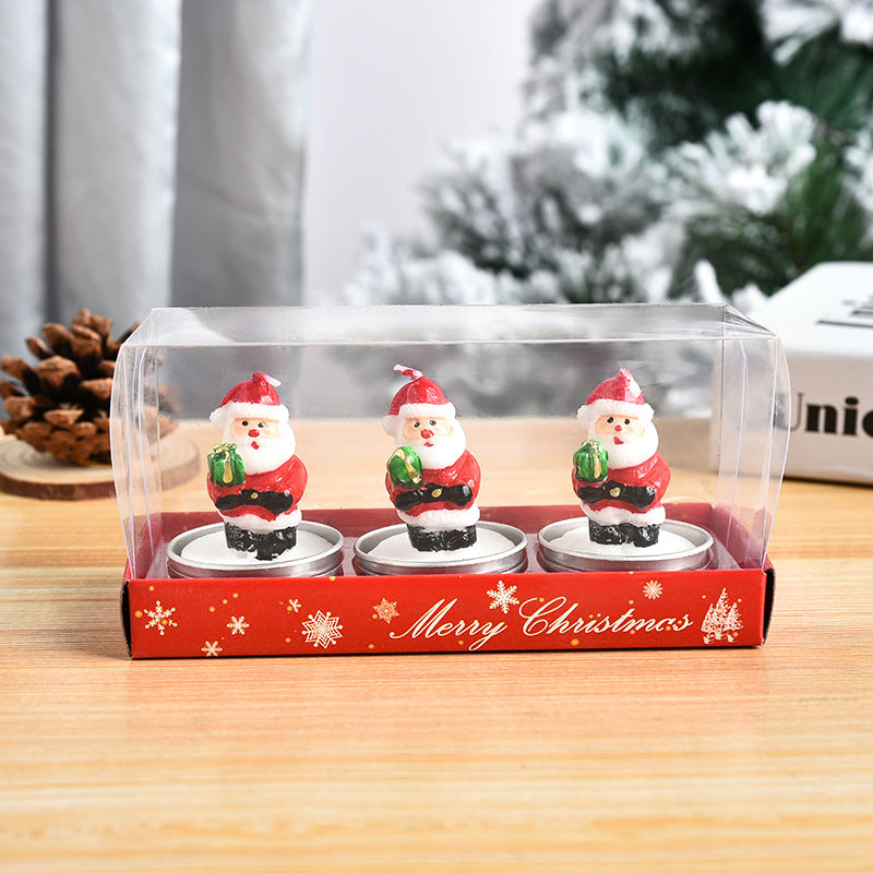 A clear box contains three Santa-shaped candles holding green gifts, set on a wooden table. The red box reads "Merry Christmas," with pine cones and a snowy backdrop.