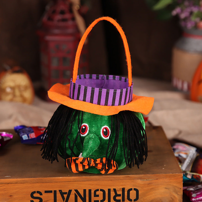 A green, plush witch basket with black yarn hair, a pointy nose, and white fangs. It features a striped purple hat with an orange brim and a matching bowtie.