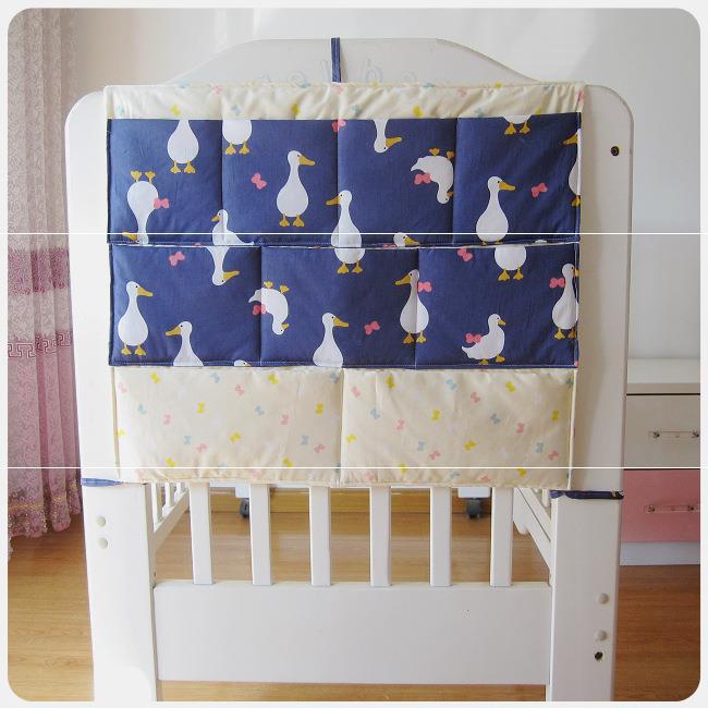 A baby crib organizer with a white and dark blue duck pattern fabric, featuring multiple pockets filled with yellow, pink, and light blue motifs. The crib is white with wooden flooring.