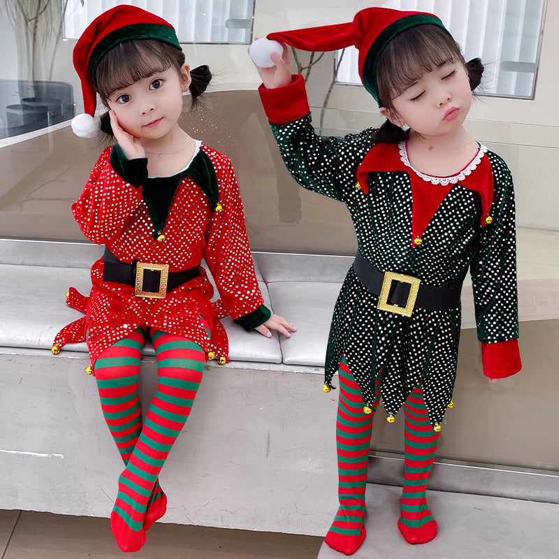 Two young children dressed as elves sit on a bench. One wears a red outfit, the other a green one, both with striped tights, Santa hats, and festive belts.