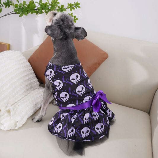 A small dog is sitting on a couch, wearing a black dress with white skull patterns and a purple bow. The background features green leaves and white cushions.
